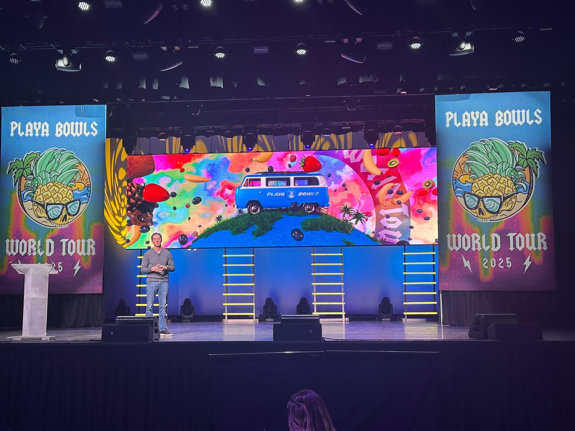 A person stands on a stage in front of a digital screen featuring a blue van and banners for 