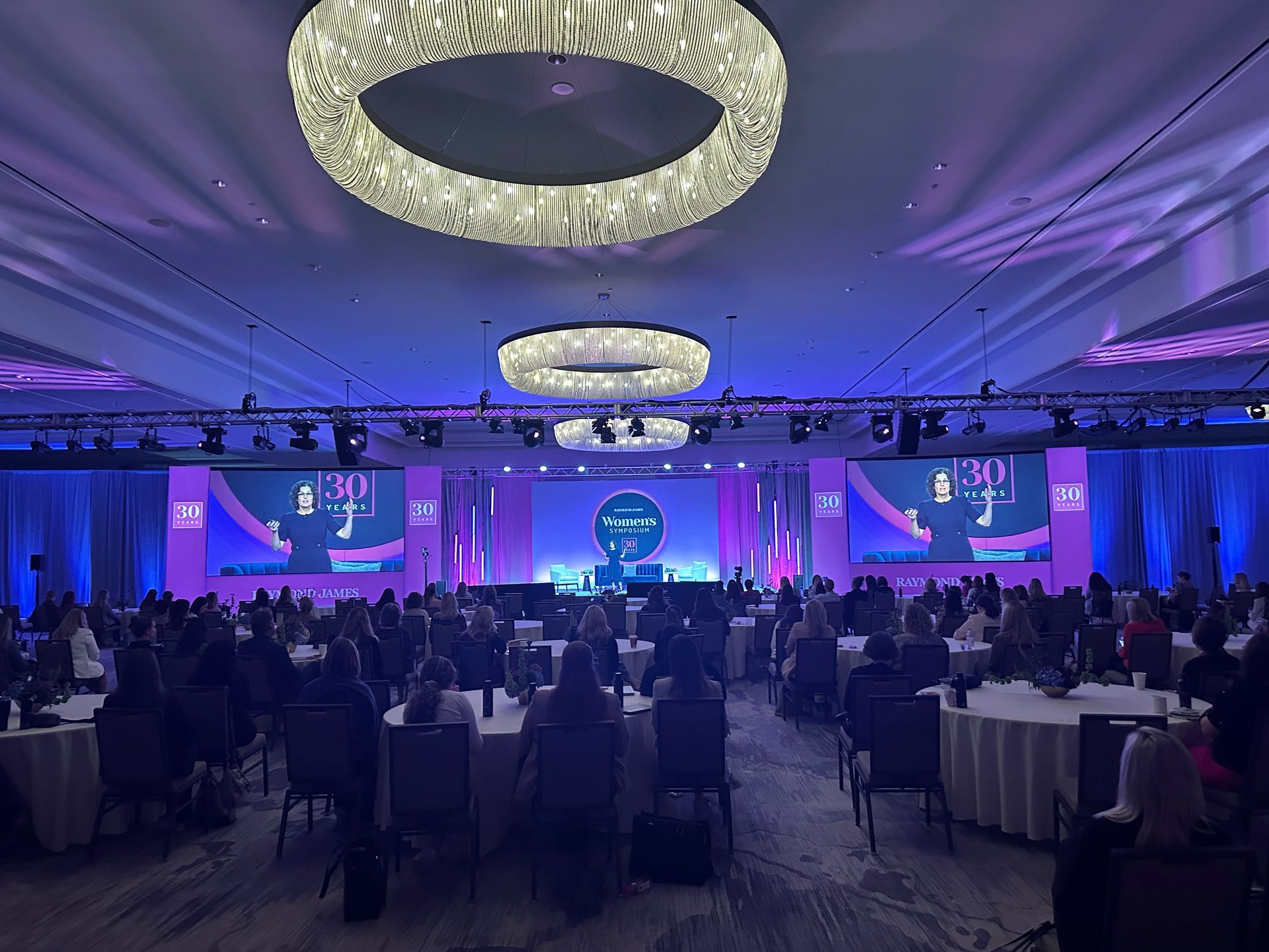 A large auditorium features a stage with digital displays, round tables with seated attendees, and ornate chandeliers.