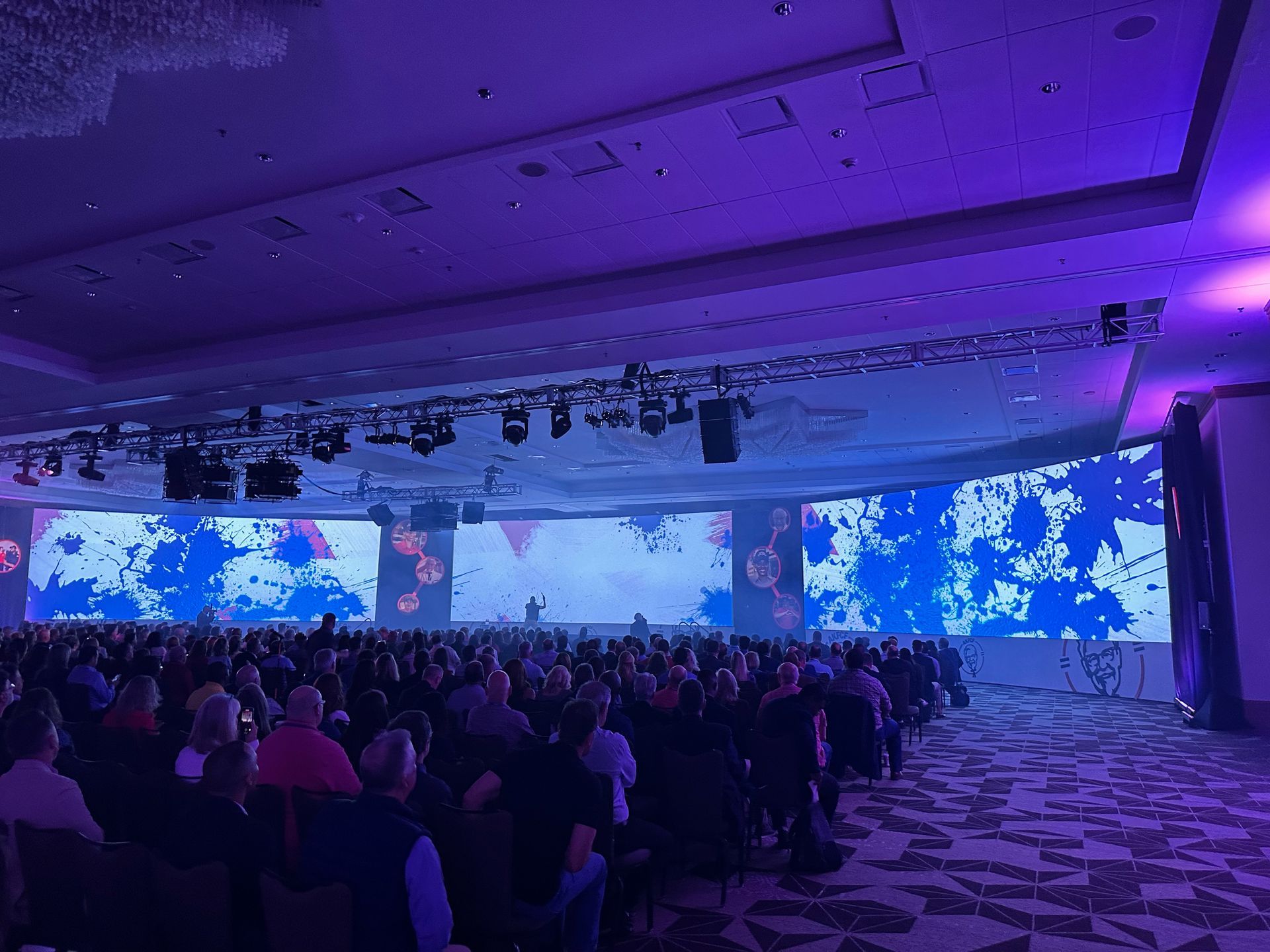 A large, dimly lit conference hall with a screen displaying blue abstract patterns, facing a seated audience.