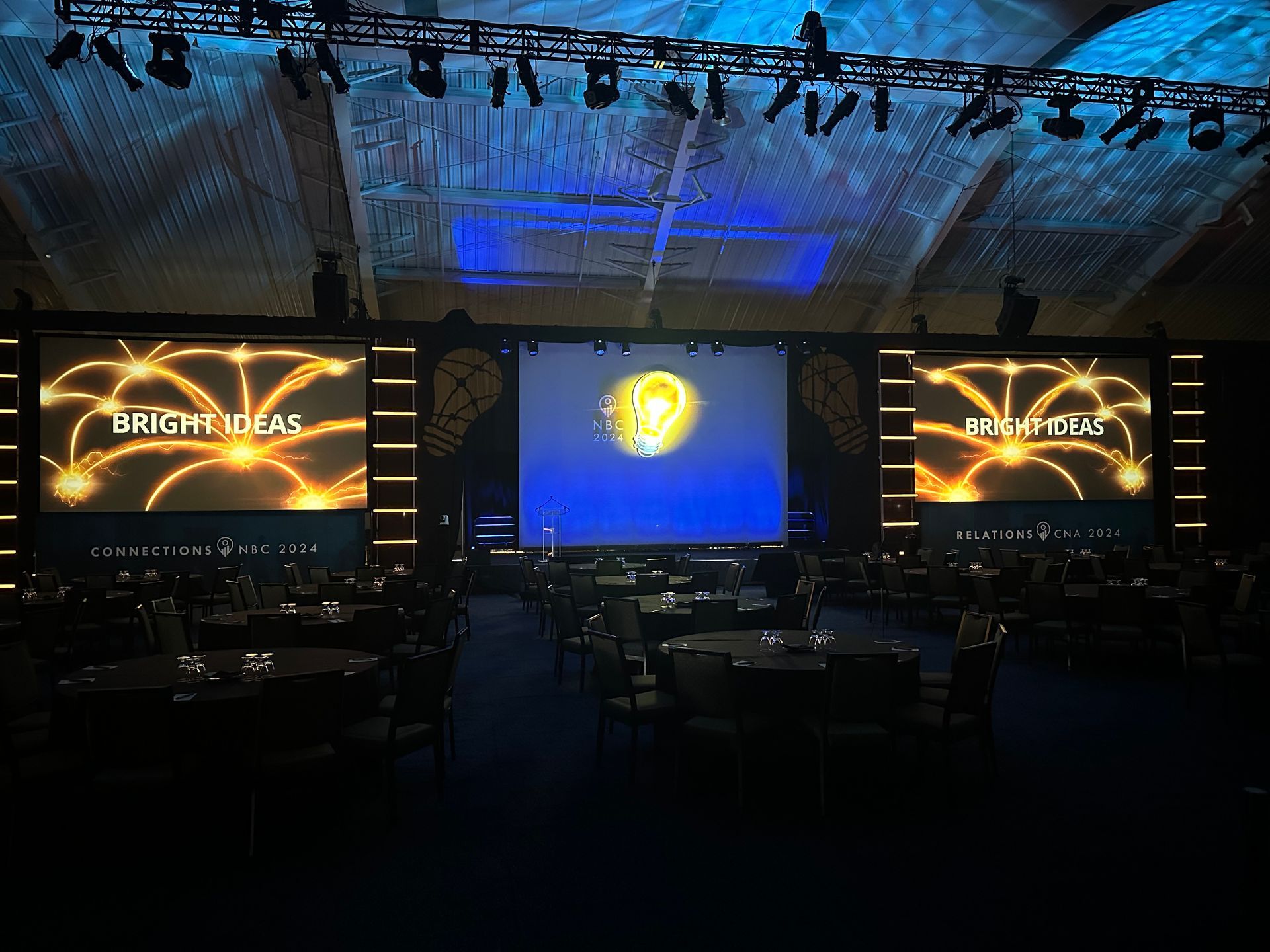 A dark event hall features three large screens displaying