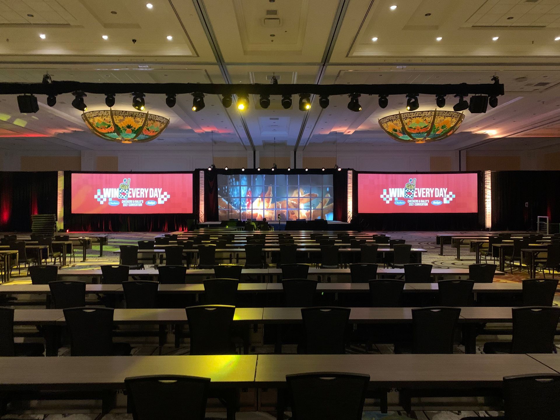 A large ballroom set up for a conference with three expansive projector screens displaying a red logo, facing rows of seats.