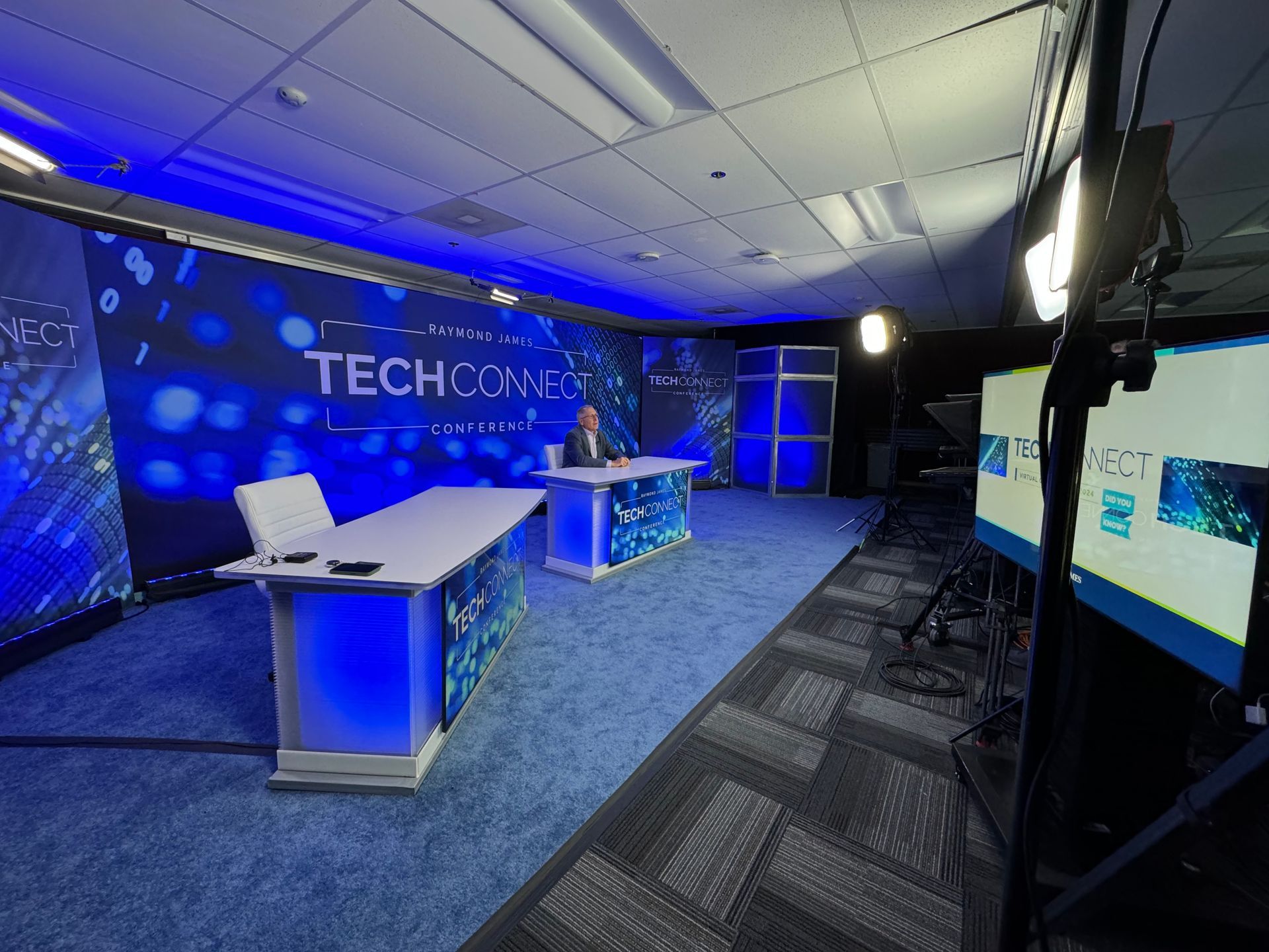 A broadcast studio set with a long desk, blue lighting, and a screen displaying the