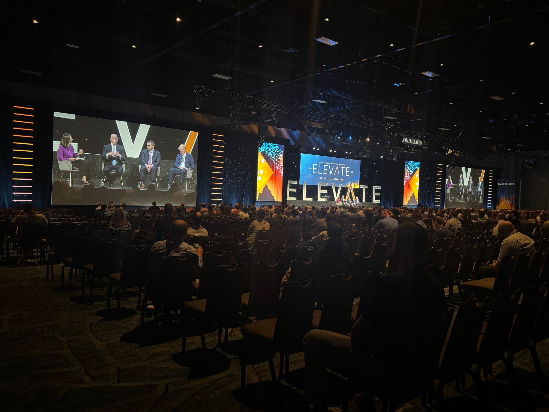 A large audience sits in a dark auditorium watching a panel discussion on a large stage screen during the Elevate event.