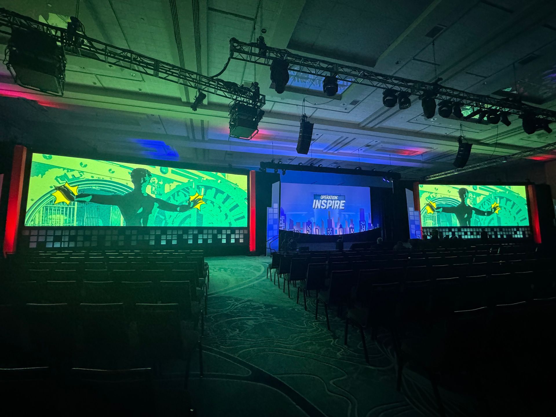 A dimly lit conference room with three large video screens showing a digital figure, set above rows of empty chairs.