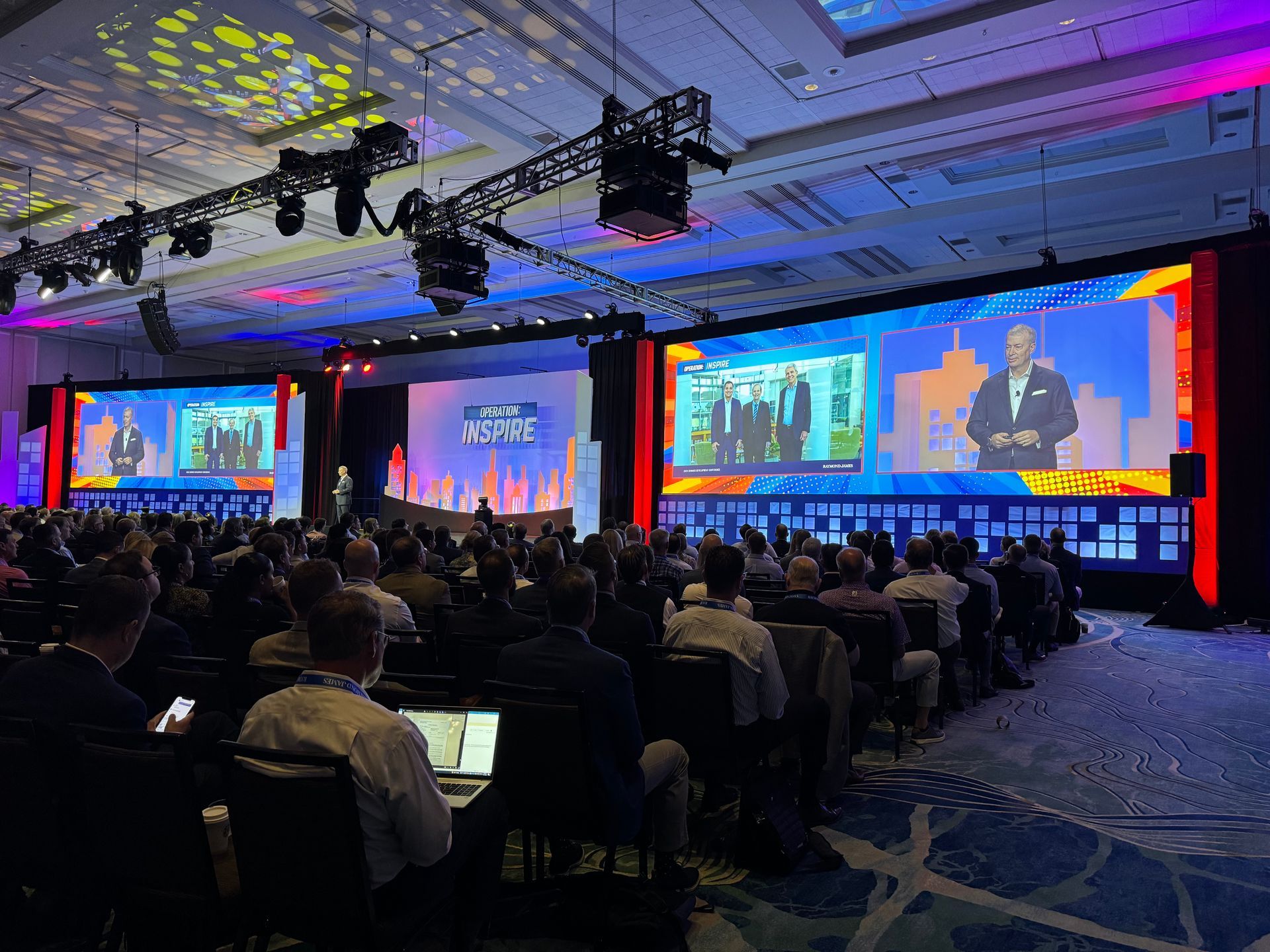 A professional conference features a large screen with a speaker, viewed by a seated audience in a dimly lit ballroom.