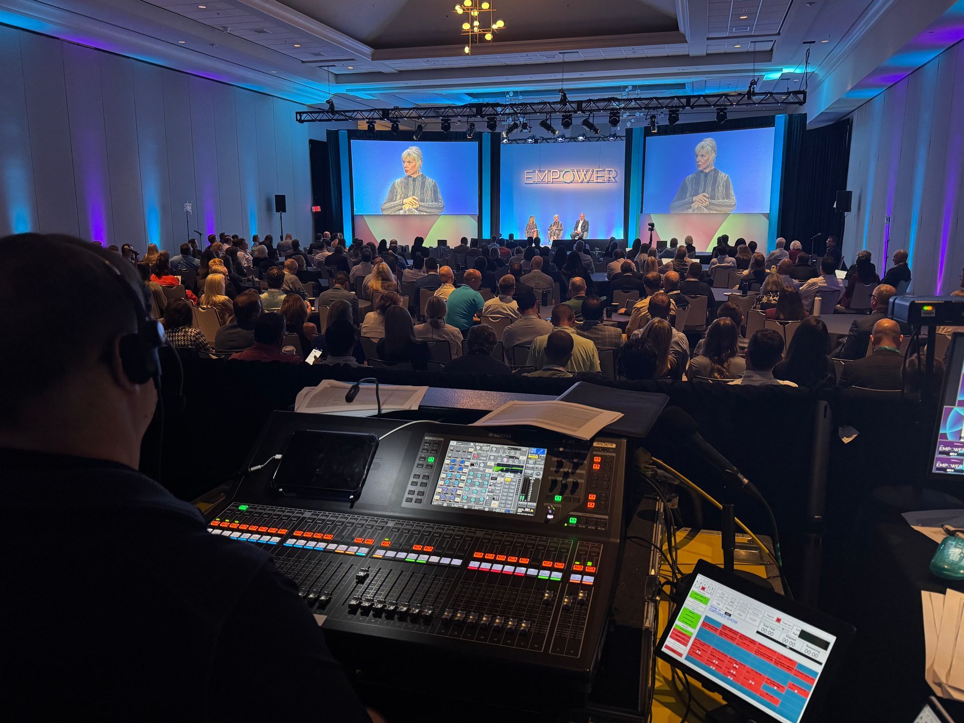 An audio technician controls a sound board at a large conference, viewing a stage with speakers and a seated audience.