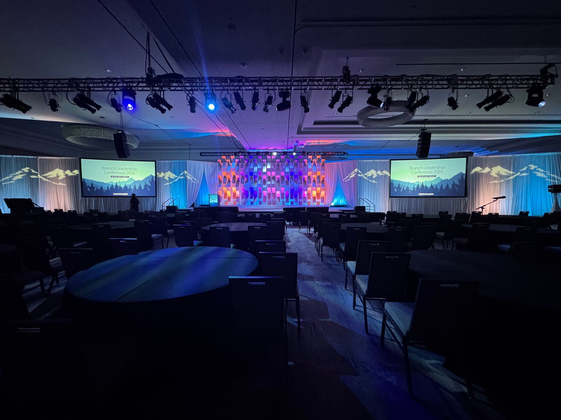 A dark event hall with a lit stage, two large screens, and circular tables arranged in a conference setup.