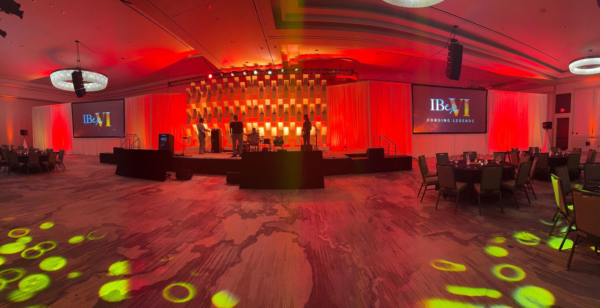 A banquet hall set up for an event, featuring a stage with a lit, geometric backdrop, red lighting, and round tables.