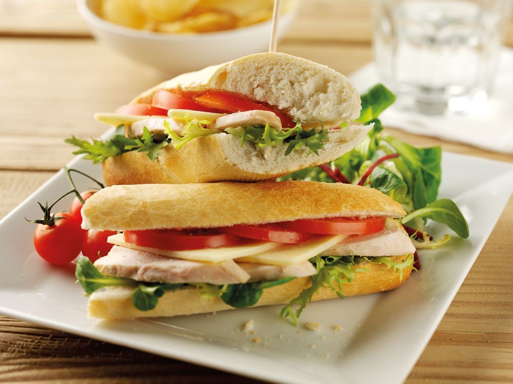 Two-tier sandwich with lettuce and tomato on a white plate, served with salad and chips.