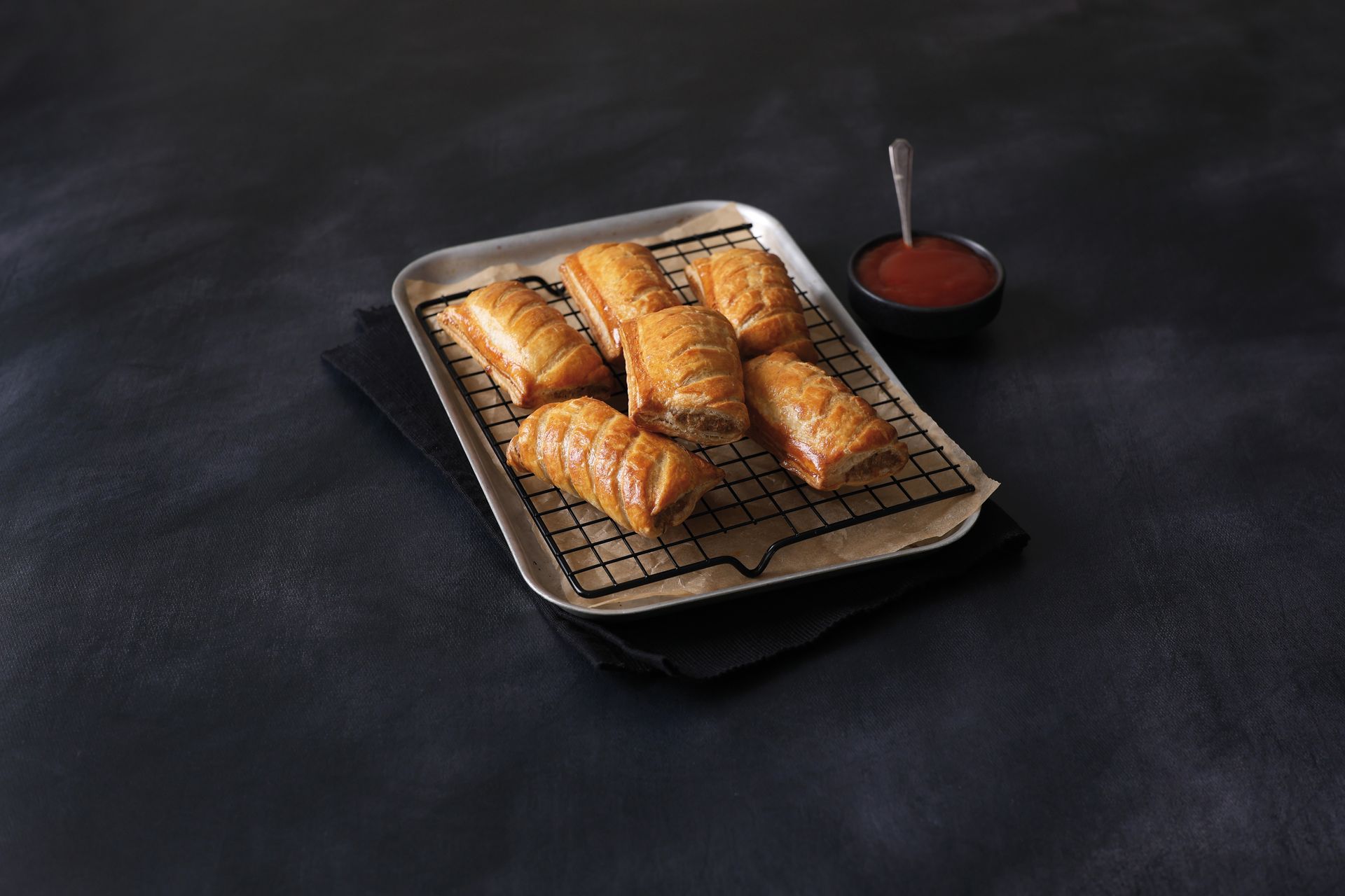 Four golden-brown pastries on a plate with a small cup of red dipping sauce on a dark table.
