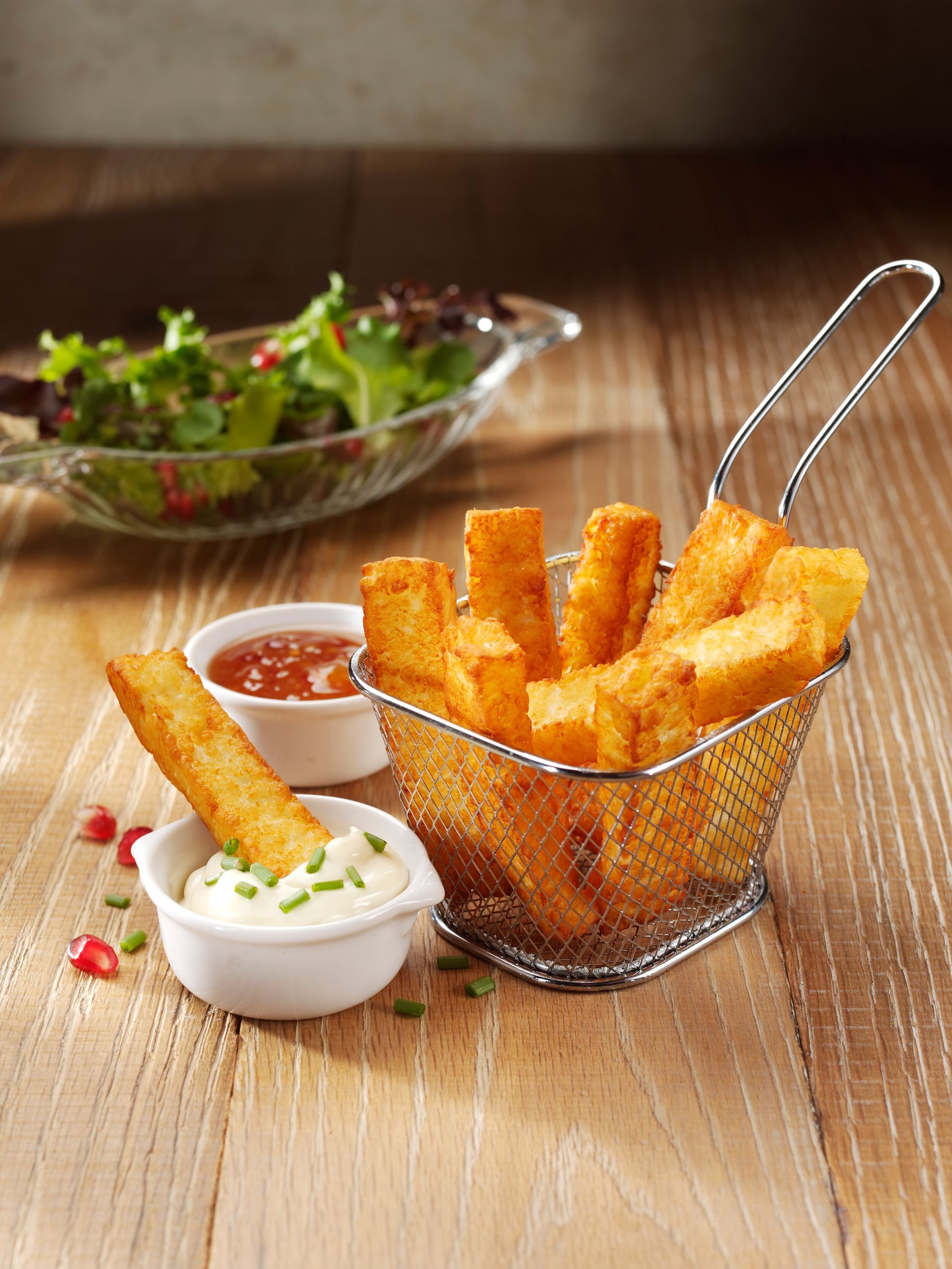 French fries in a wire basket with dipping sauces on a wooden table, with a salad in the background