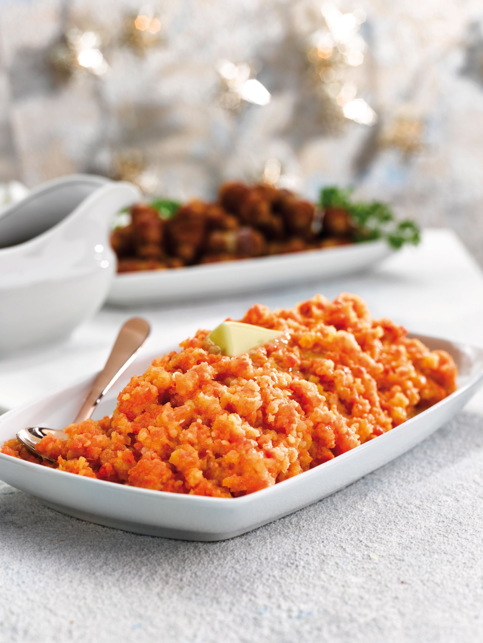 Orange couscous in a white serving dish with butter, set on a light table with another dish blurred behind