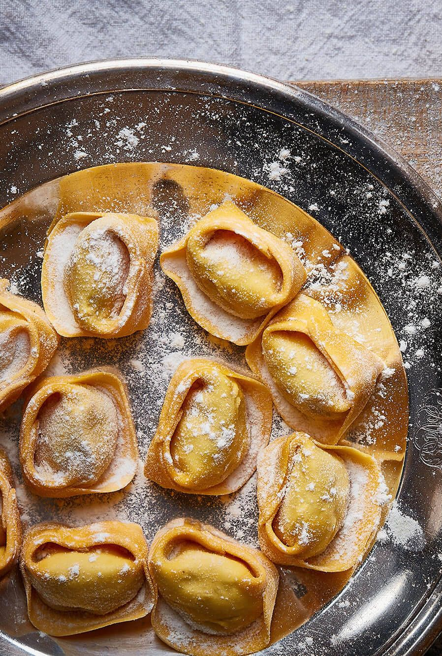 Homemade ravioli on a dark plate, dusted with flour, beside a wooden pasta wheel on a rustic surface