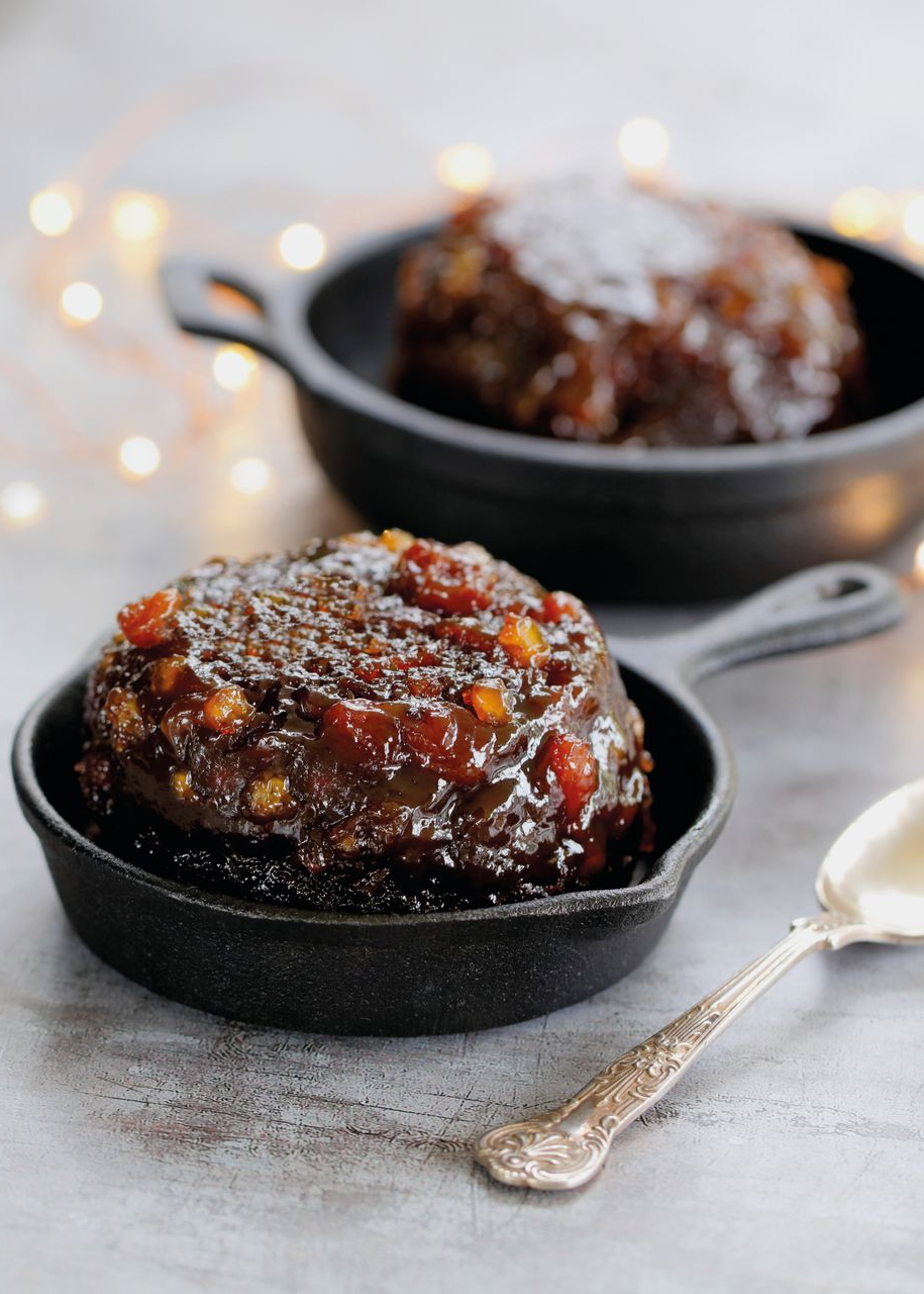Two glazed chocolate desserts in black dishes with a spoon on a light tabletop, softly lit with fairy lights