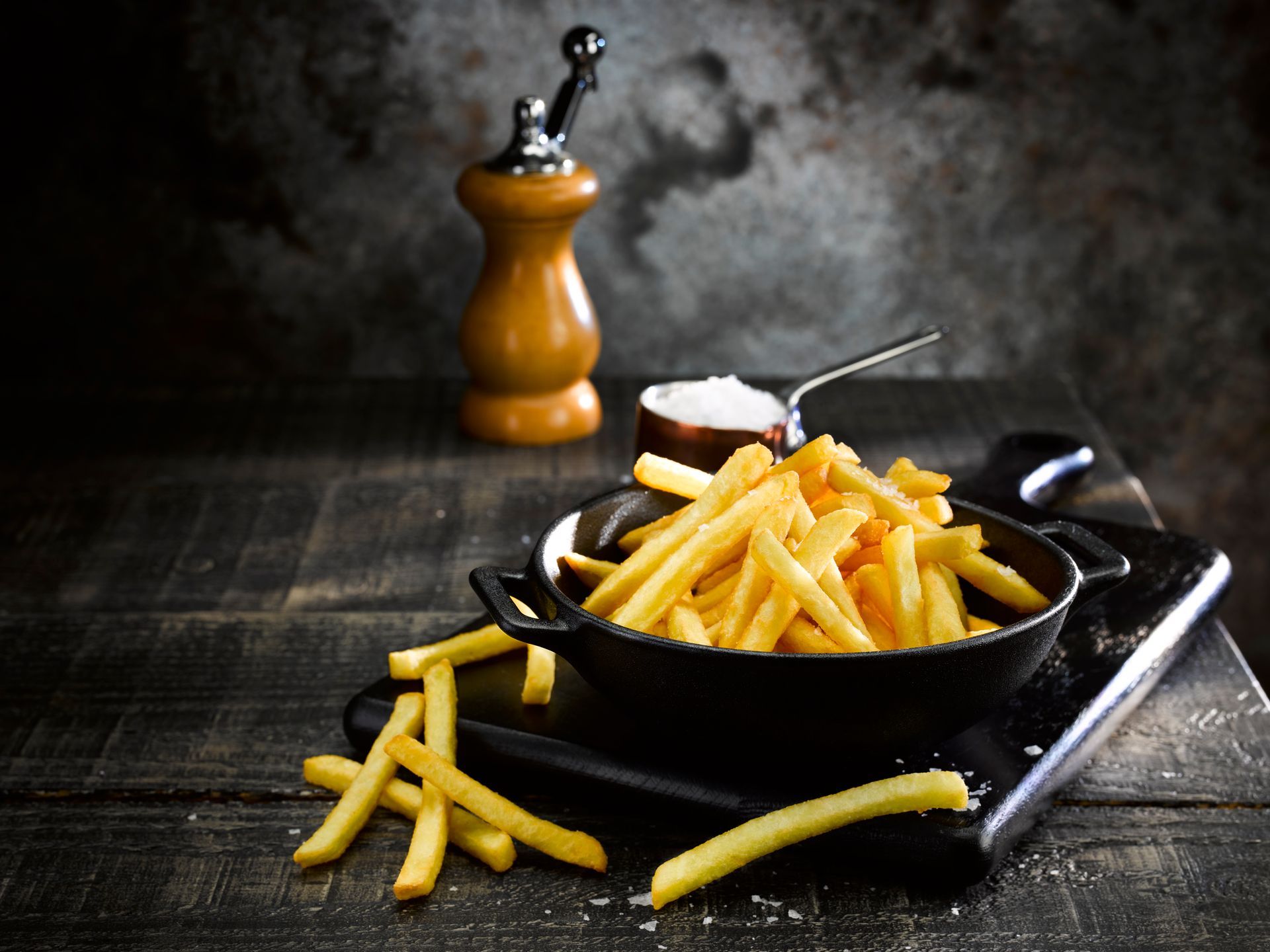 French fries in a black pan with a wooden pepper grinder on a dark table