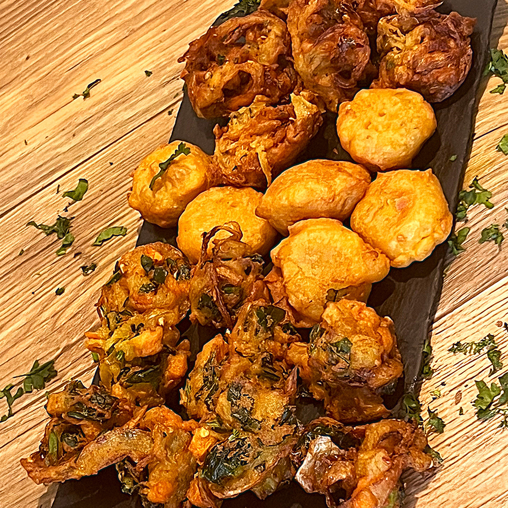 Tray of golden-brown fried snacks and pakoras on a wooden table, garnished with chopped herbs