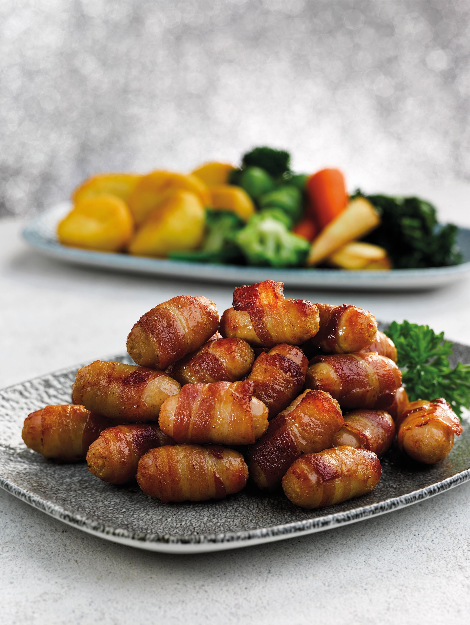 Plate of glazed bacon-wrapped bites on a table, with a blurred platter of vegetables in the background