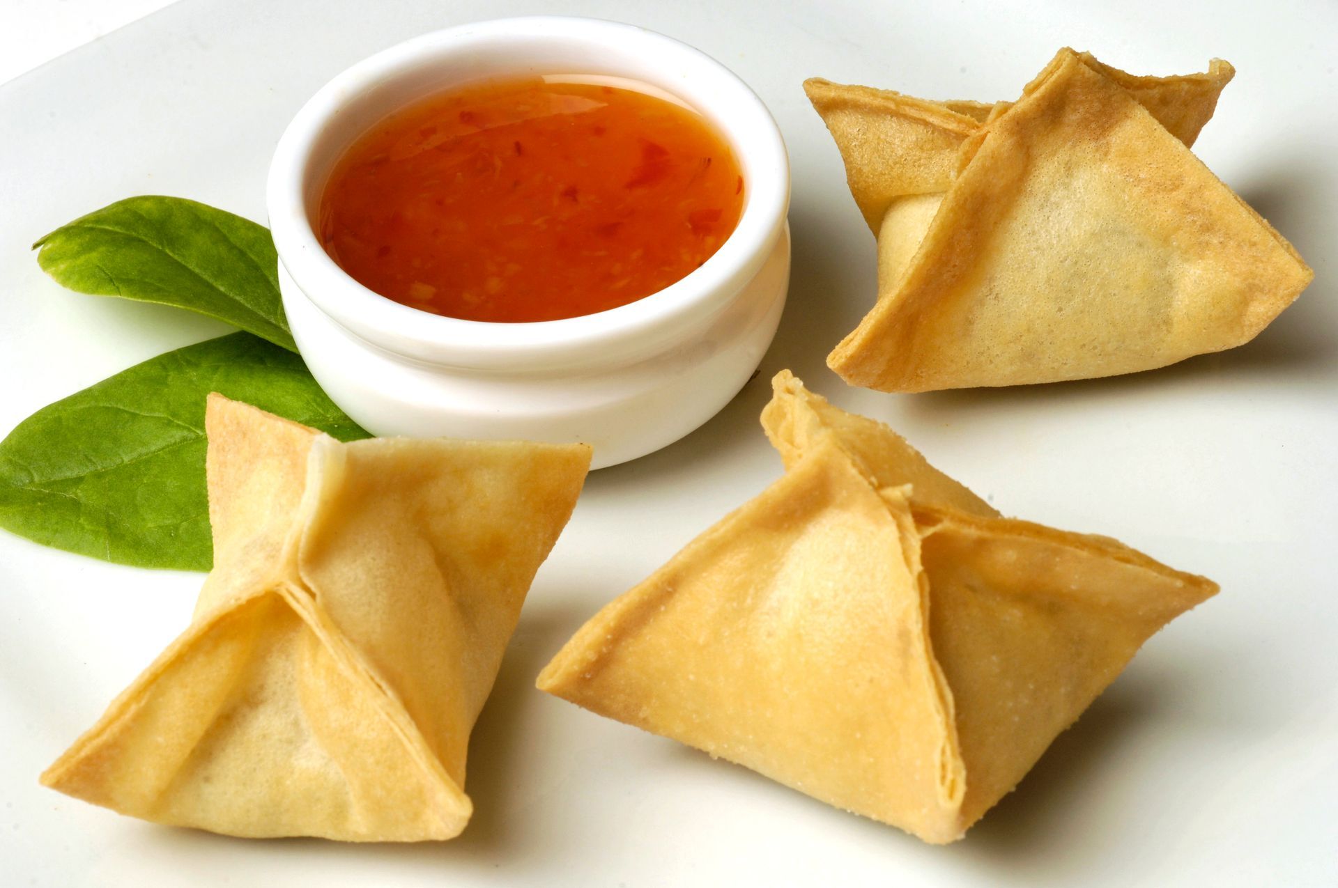 Fried wonton triangles with red dipping sauce and green leaves on a white background