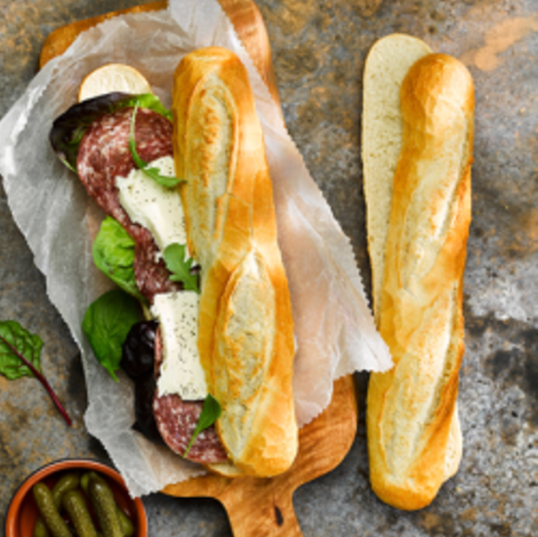Baguette sandwich with salami, greens, and cheese beside a plain baguette on parchment