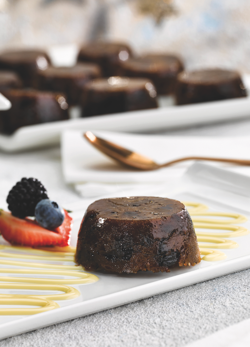 Chocolate dessert on a white plate with berry garnish and sauce drizzle, with more cakes blurred in the background