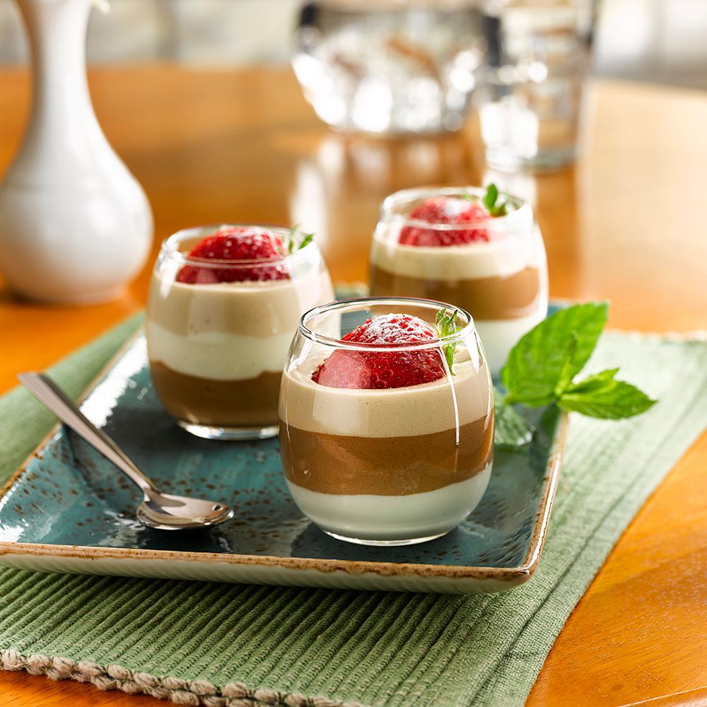 Three layered dessert cups topped with strawberries on a green placemat, with a spoon beside them