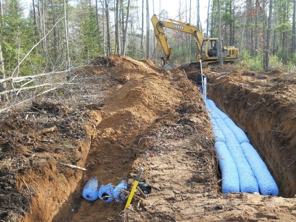 Excavator beside a trench with blue pipes being laid through a forested construction site