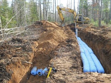 Excavator trenching in a wooded site with blue pipes laid in the ditch