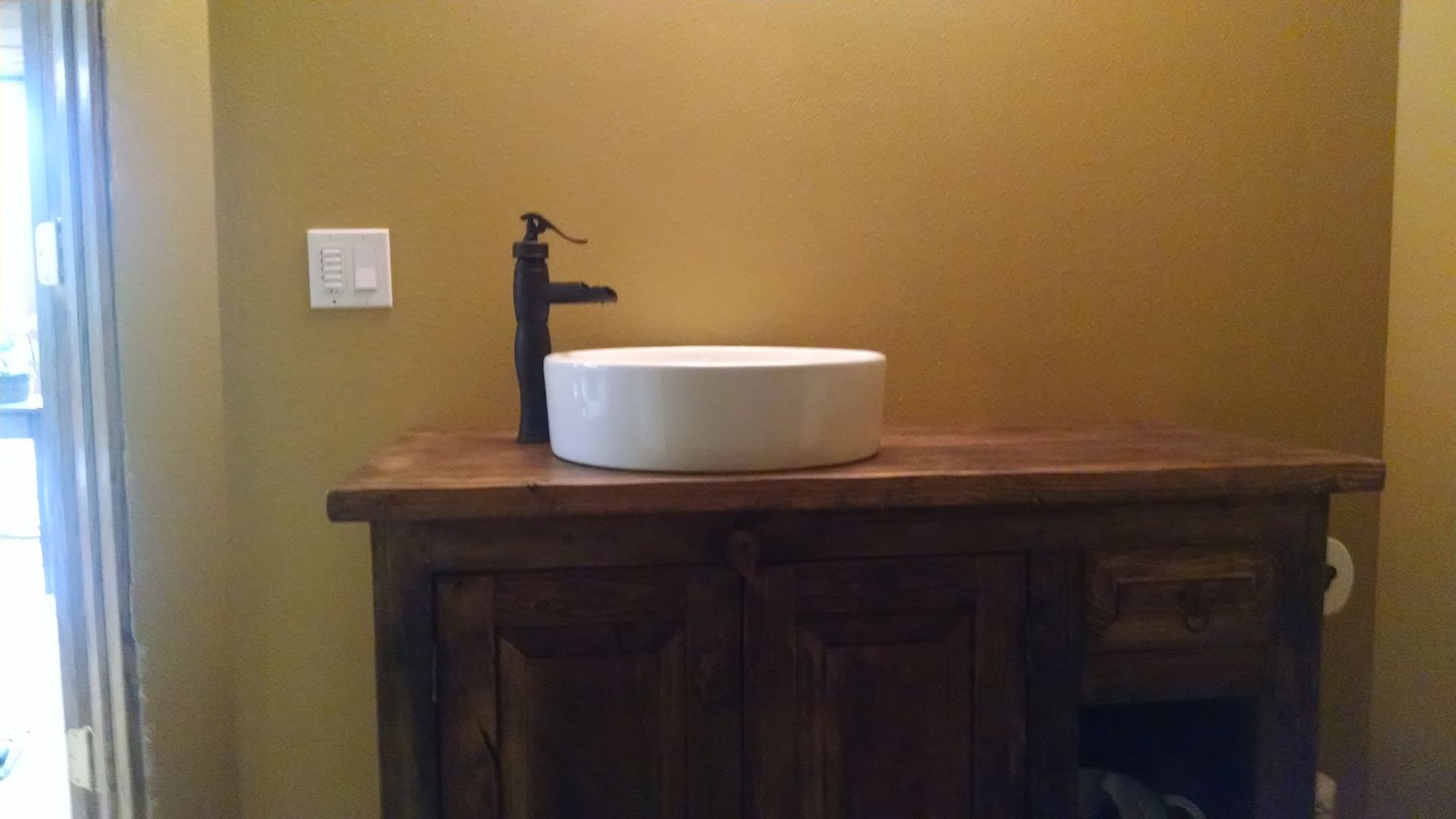 Bathroom vanity with a white vessel sink and dark faucet on a wooden cabinet against a tan wall.