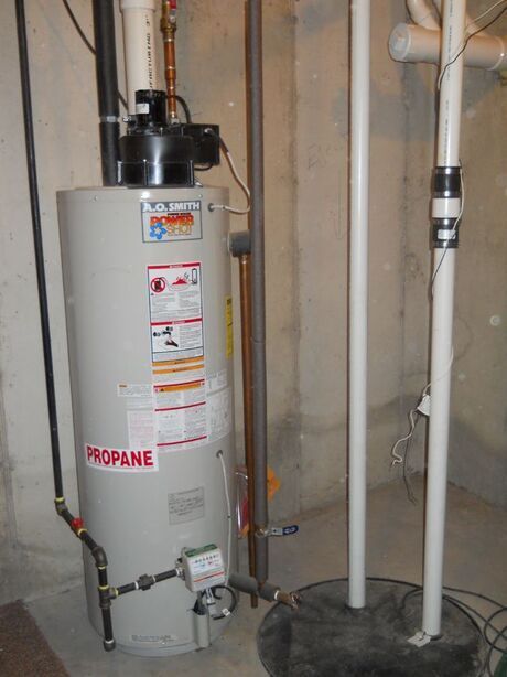 Gray propane water heater in a basement, with exposed pipes and utility lines nearby.