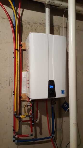 White wall-mounted boiler with blue display in utility room, surrounded by exposed red, yellow, and white pipes.