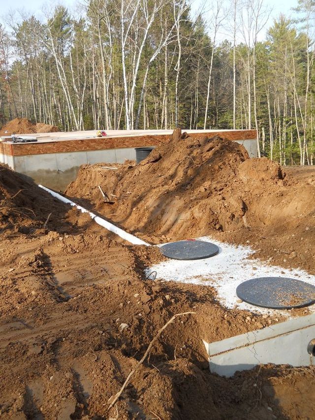 Construction site with excavated dirt, concrete foundation, and two round access covers near a wooded area
