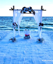White beach wedding arch with blue flowers on the sand by the ocean at sunset