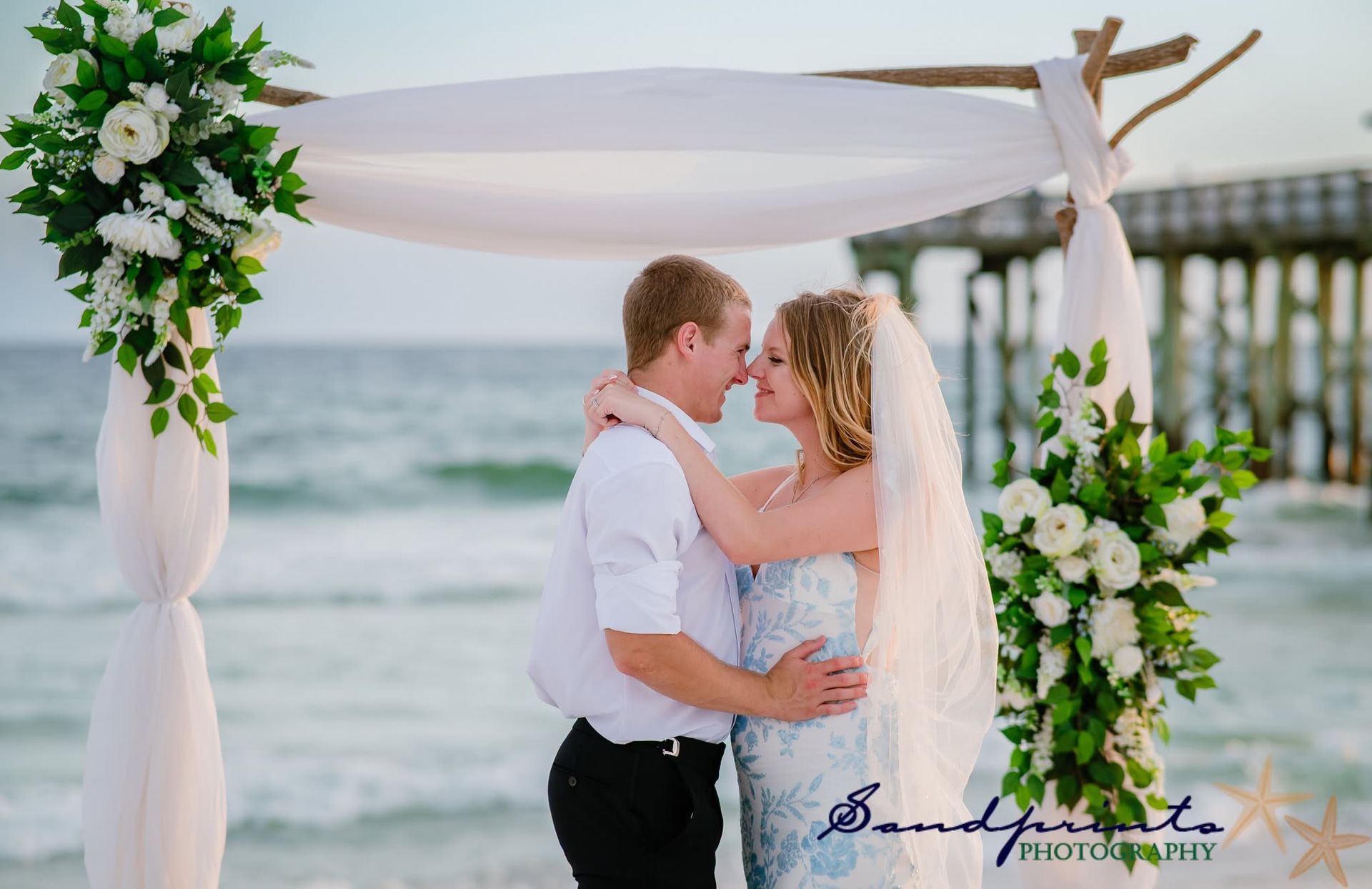 Couple embracing under a white beach wedding arch with floral arrangements by the ocean