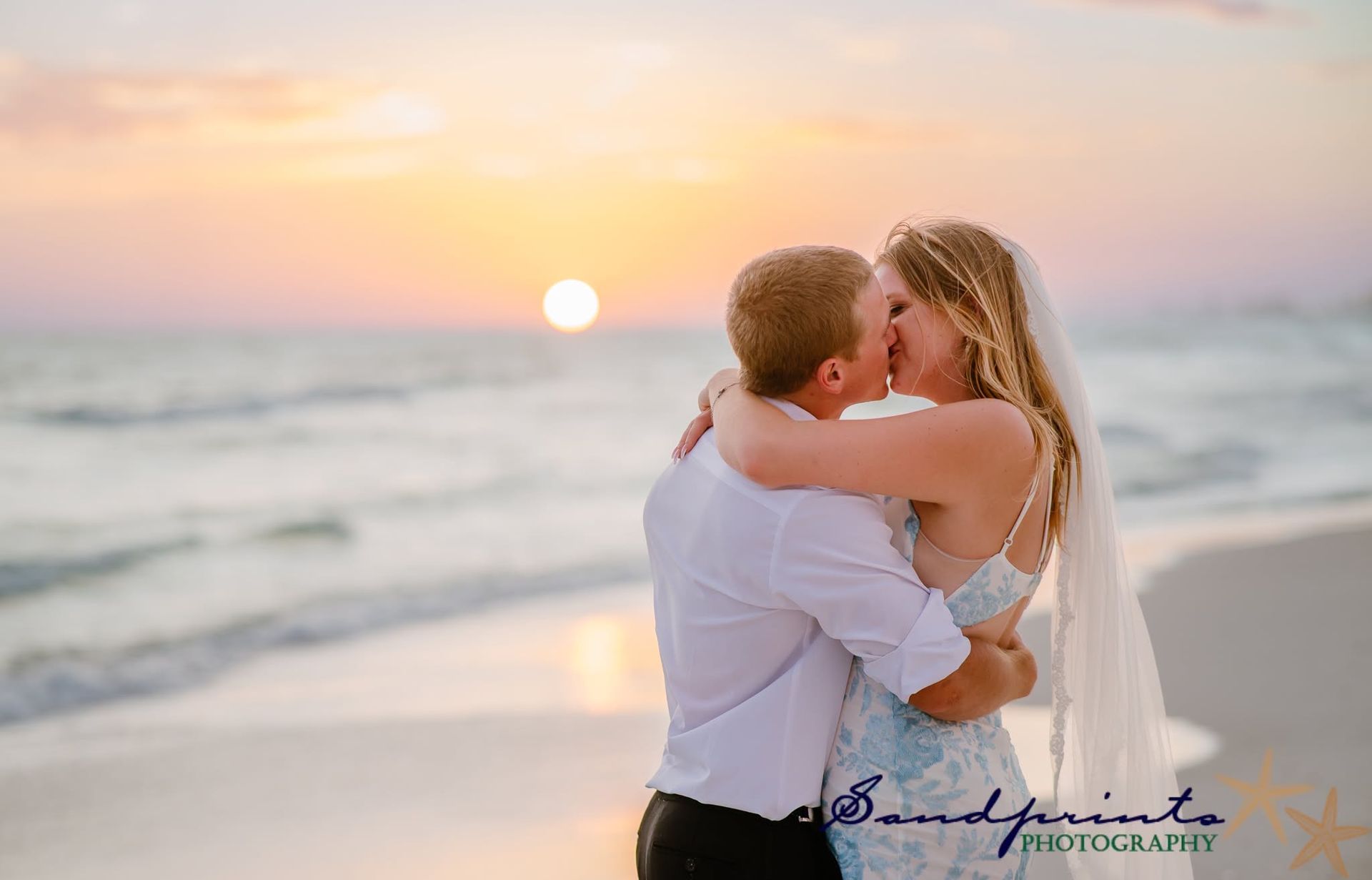 Couple embracing and kissing on a beach at sunset, with waves and warm pastel sky in the background