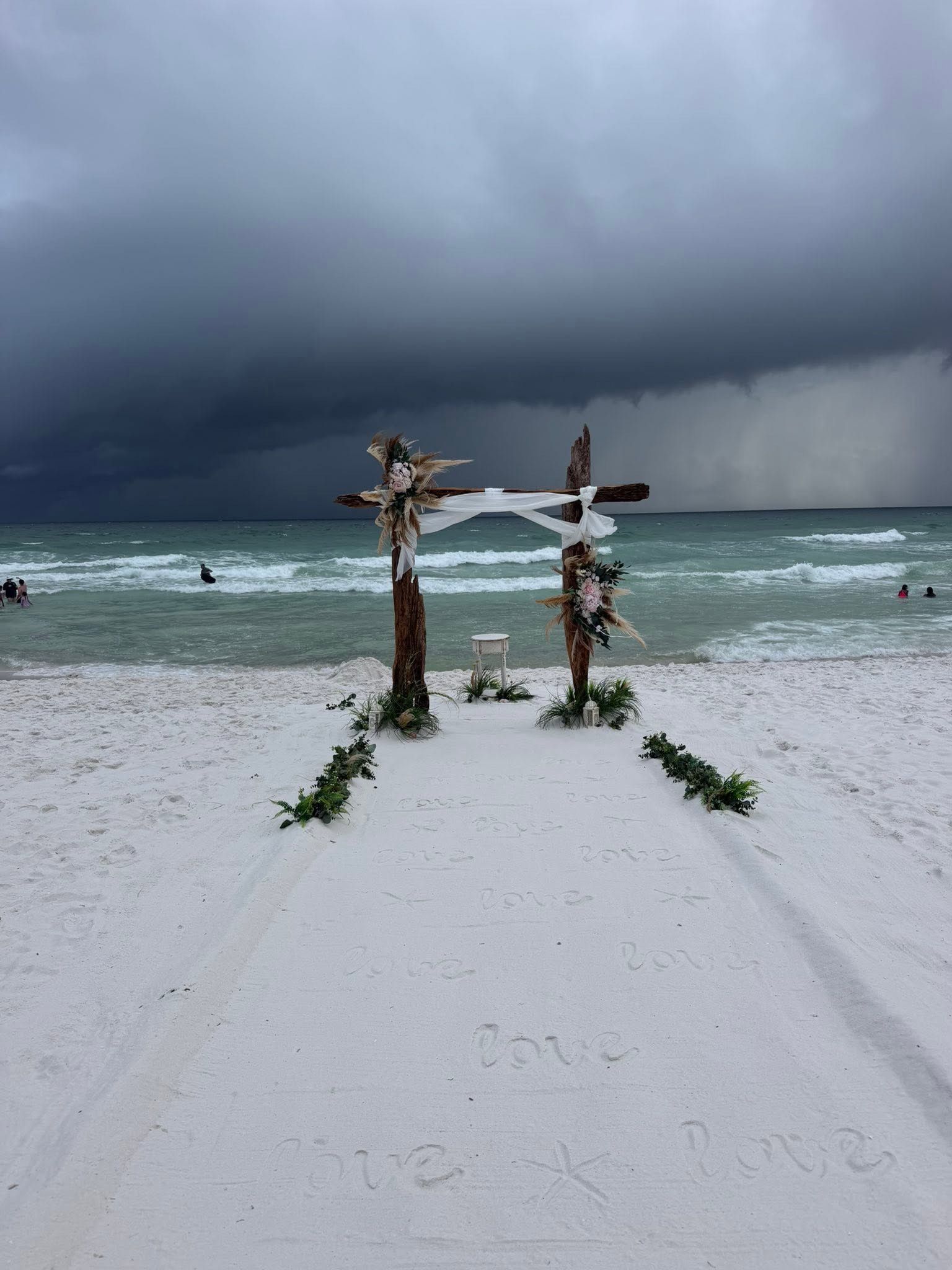 Storm clouds over a white-sand beach with two wooden posts and distant swimmers in turquoise water