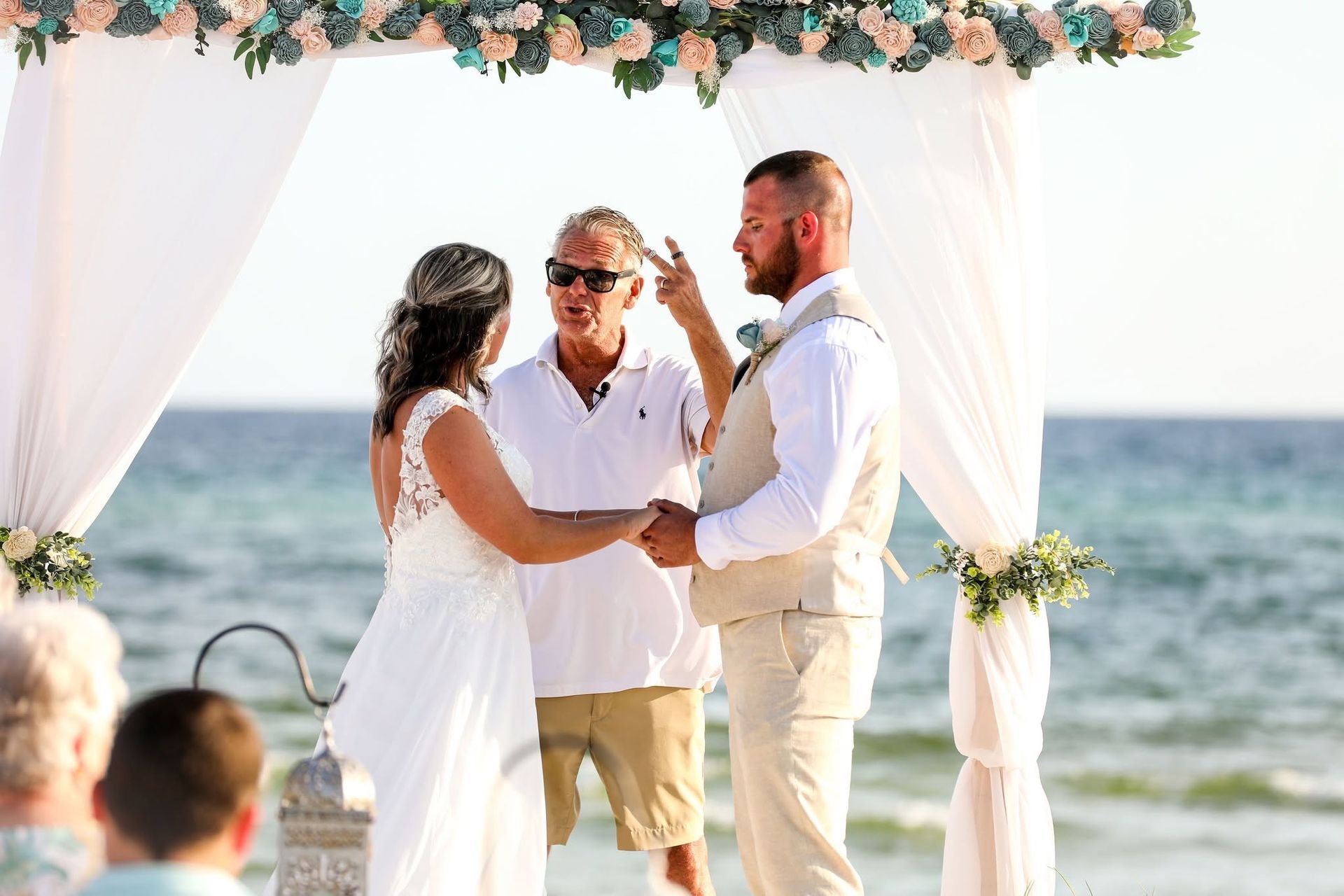 Beach wedding ceremony under a flower-decorated arch, with an officiant and couple holding hands by the sea.
