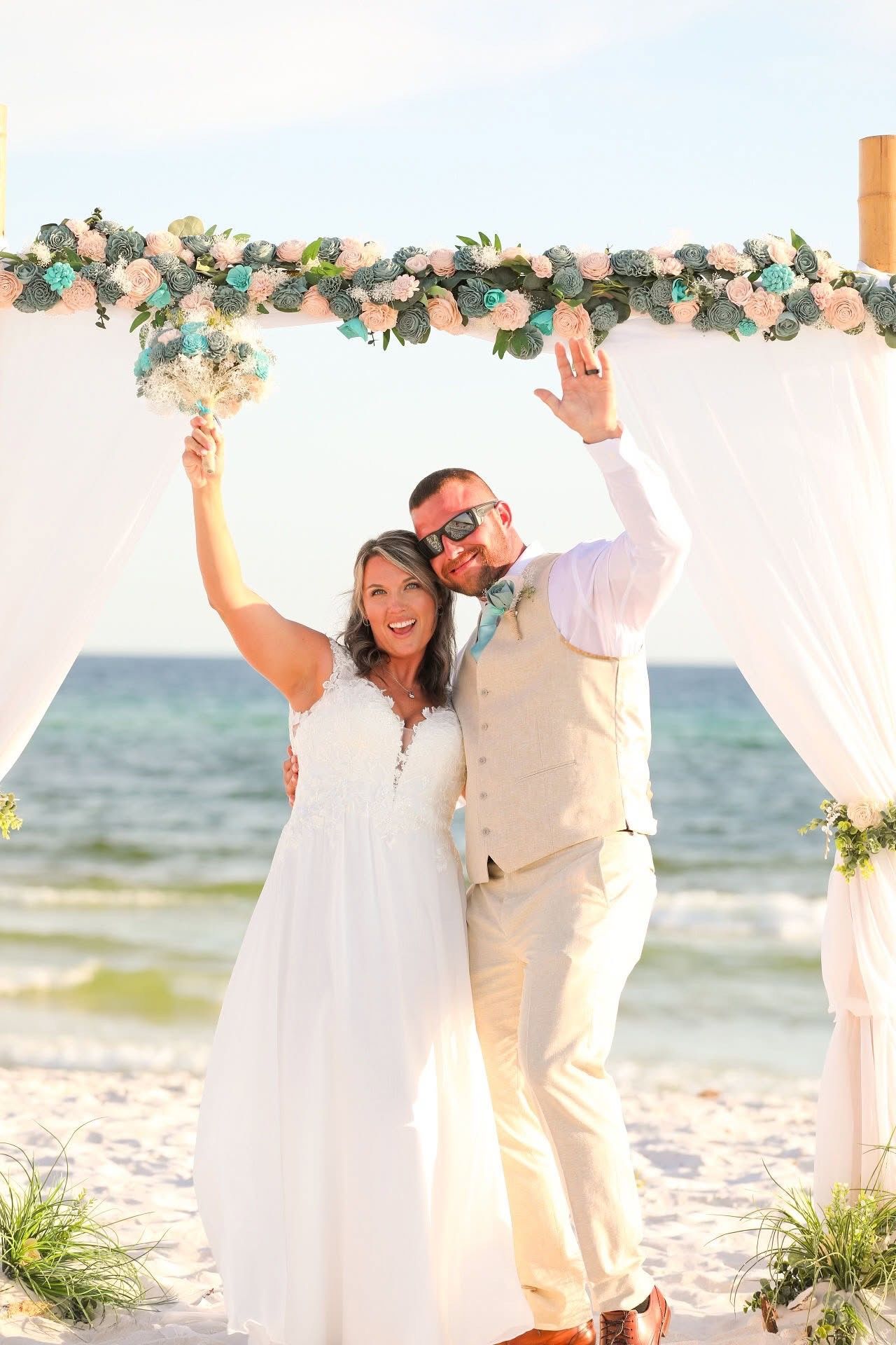 Couple in white attire cheering under a flowered arch on a sunny beach ceremony platform