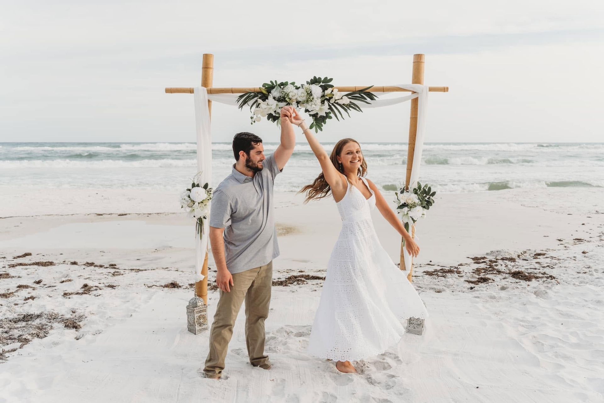 Couple holding hands under a decorated arch on a beach with waves in the background
