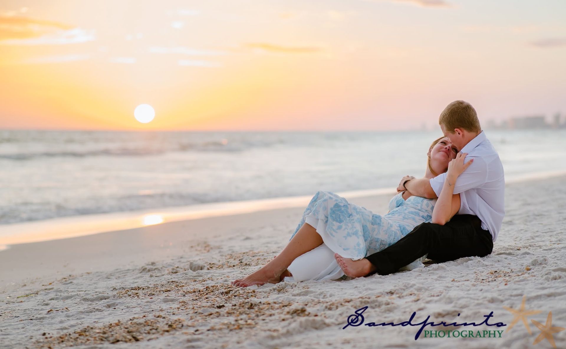 Couple embracing on a sandy beach at sunset, with gentle waves and pastel sky in the background.