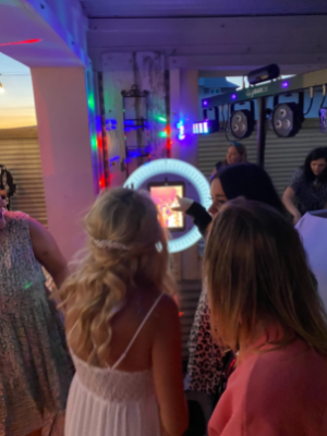 People viewing a glowing ring light booth at a lively party, with colorful lights and hanging decorations.