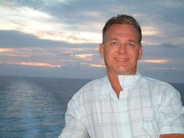 Smiling man in a white plaid shirt on a boat at sunset, with the ocean behind him