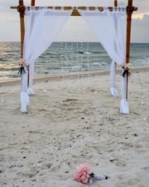 White wedding arch with draped curtains on a sandy beach beside the ocean