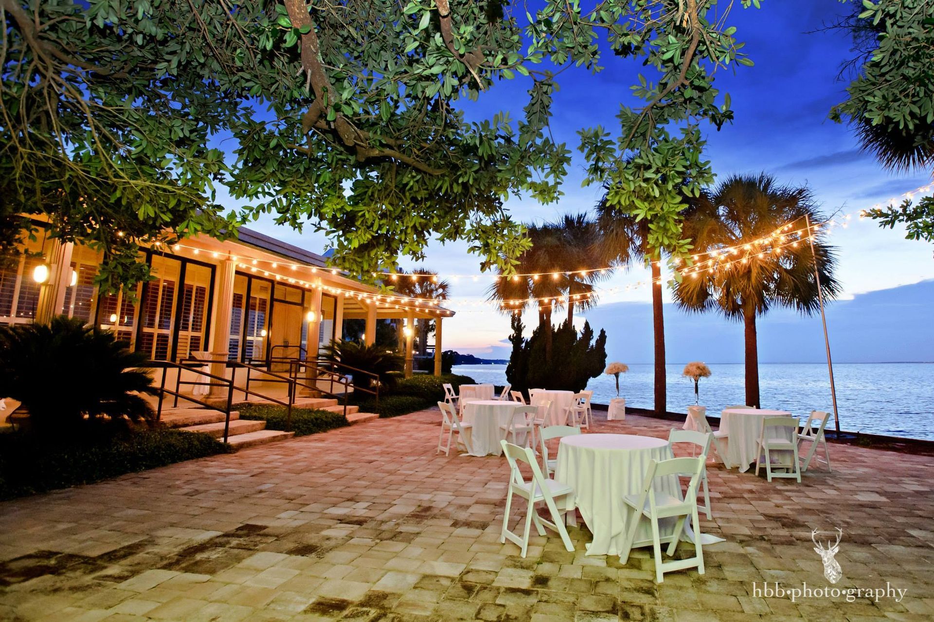 Outdoor seaside patio with white tables under string lights beside a lit building at dusk