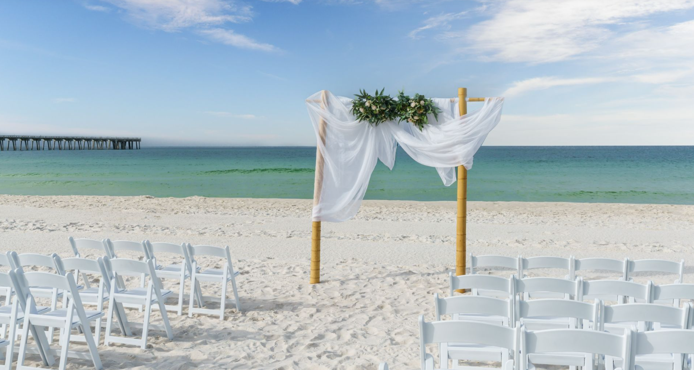 Beach wedding arch with white drapes and flowers, flanked by rows of white chairs on a sandy shore