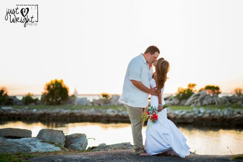Couple embracing by a lakeside at sunset, with flowers and a rocky shoreline.