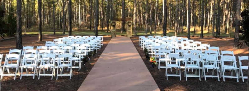 Outdoor wedding ceremony setup with white chairs lining a wooden aisle in a wooded grove