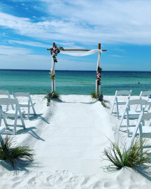 Beach wedding aisle with white chairs and floral arch facing the ocean under a blue sky