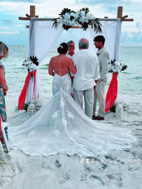 Beach wedding ceremony under a white floral arch, with bride and groom facing the officiant at sunset.