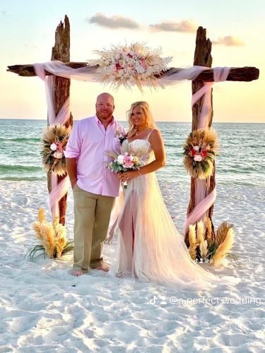 Couple standing on a decorated beach wedding arch at sunset, holding bouquets with the ocean behind them