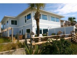 White two-story house with a wooden deck, fenced yard, and palm tree under a clear blue sky